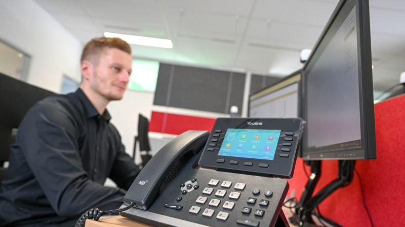 Zu sehen ist ein Telefon mit Eingabemodul. Dahinter sitzt ein blonder Mann, der auf zwei Bildschirme blickt.