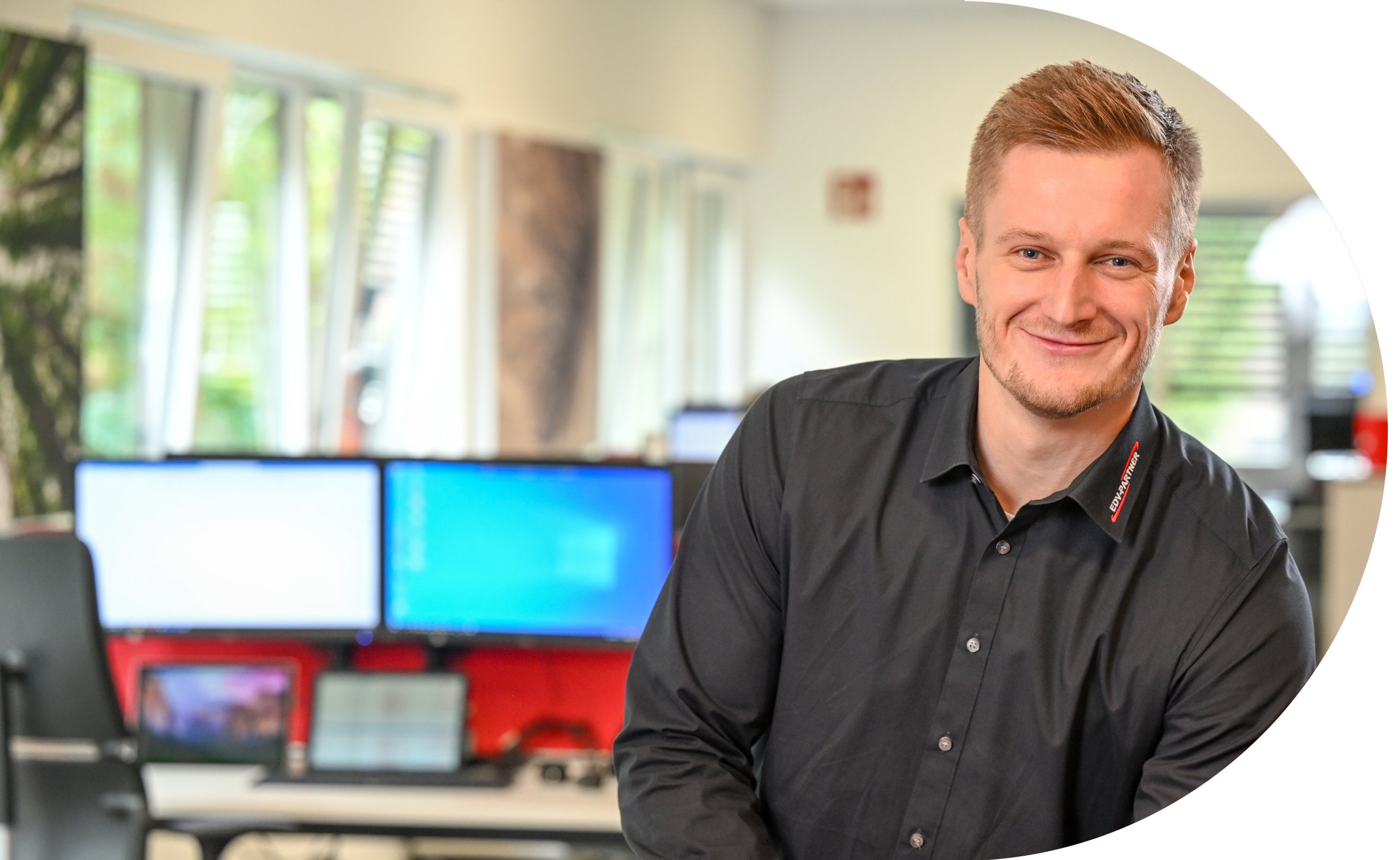 Ein Mann mit blonden Haaren Stützt sich auf etwas und lächelt freundlich in die Kamera. Im Hintergrund sind mehrere eingeschaltete Bildschirme zu sehen