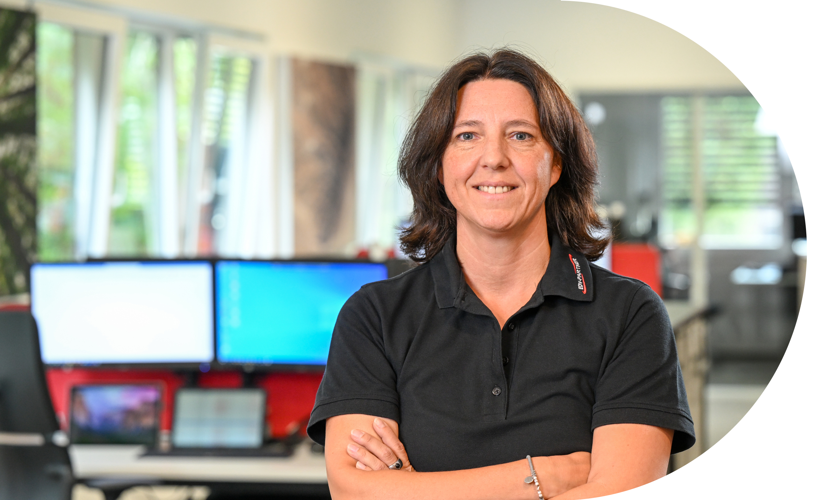 Eine Frau mit dunkelbraunen Haaren verschränkt ihre Arme und lächelt in die Kamera. Im Hintergrund sind mehrere eingeschaltete Bildschirme zu sehen.