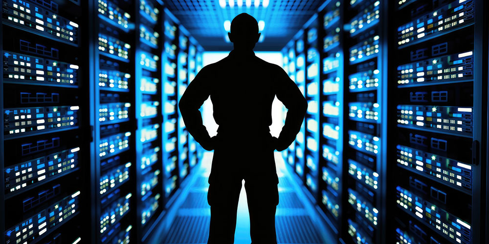 Die Silhouette eines Mannes steht in der Mitte eines langen Korridors. Zu beiden Seiten sind beleuchtete Serverschränke zu sehen.