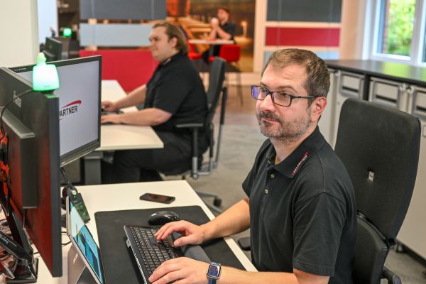 Ein Mann arbeitet an einem Computer mit drei Bildschirmen. Im Hintergrund ist ein weiterer Mitarbeiter zu sehen.