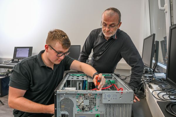 Ein IT-Technik-Azubi schraubt an einem Rechner, während der Lehrmeister ihn beobachtet.