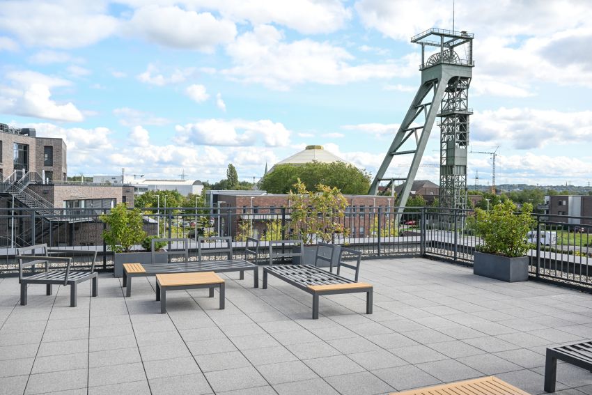 Dachterrasse mit Sitzgelegenheiten und Pflanzen, im Hintergrund ein Förderturm und städtische Gebäude bei leicht bewölktem Himmel.