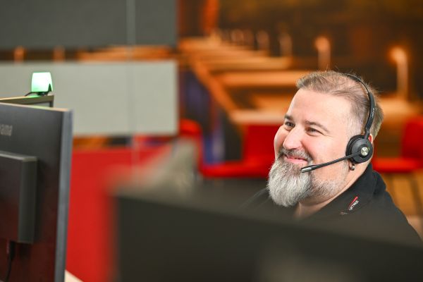 Ein Mitarbeiter lächelt freundlich, während er auf den Monitor blickt. Er trägt ein Headset und scheint sich in einem Gespräch zu befinden.