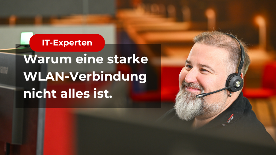 Ein Mitarbeiter lächelt freundlich, während er auf den Monitor blickt. Er trägt ein Headset und scheint sich in einem Gespräch zu befinden. Im Vordergrund ist ein Schriftzug "Warum eine starke WLAN-Verbindung nicht alles ist."