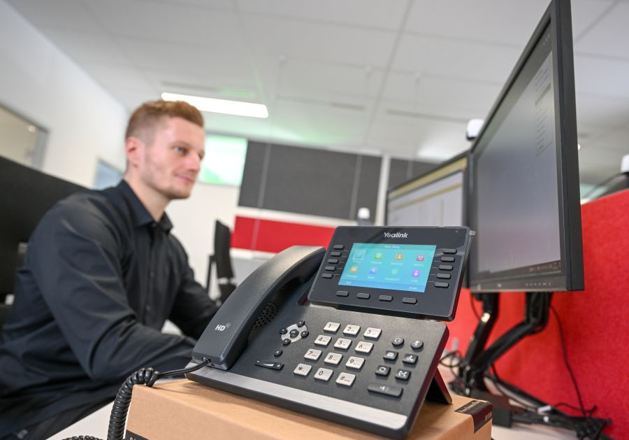 Zu sehen ist ein Telefon mit Eingabemodul. Dahinter sitzt ein blonder Mann, der auf zwei Bildschirme blickt.